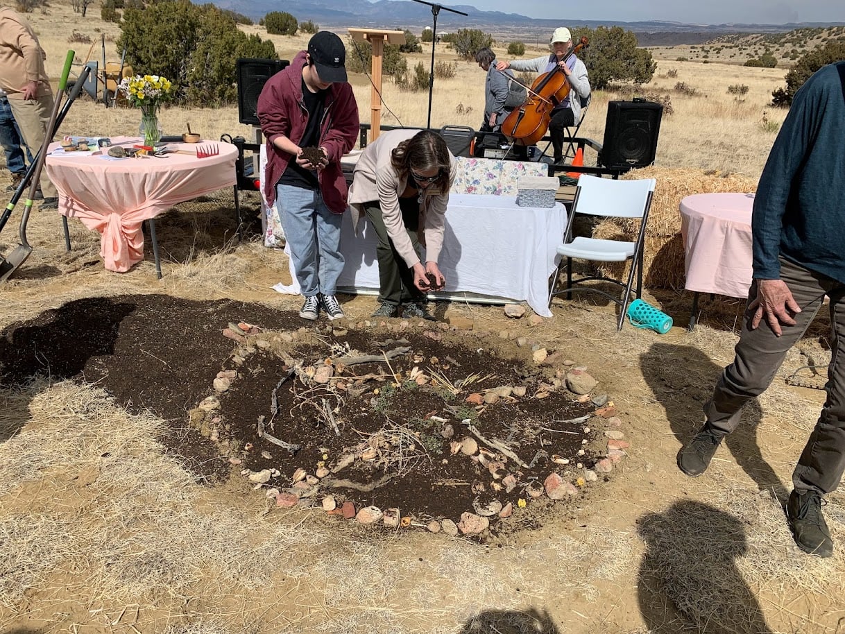 Two people placing soil on a rock-ringed earthen mound while a cellist plays behind them